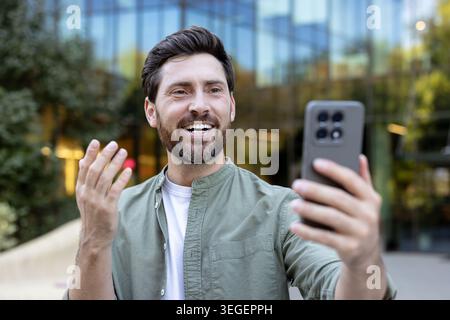 Bearded man smiling and gesturing during an outdoor smartphone video call in a downtown setting, using modern mobile technology for remote communication and connection Stock Photo