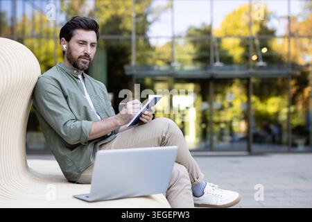 Young man with earbuds writing in a notebook while using a laptop on a modern bench in the city, focused on remote work or studying outdoors during daytime Stock Photo