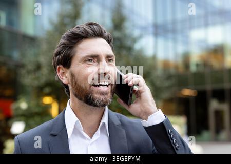 Confident businessman connecting with a client or colleague, discussing business on a cell phone, standing outside a modern office building, symbolizing success and professional communication Stock Photo