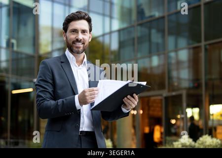 Smiling businessman reviewing business documents on a clipboard, standing in front of a modern glass office building, symbolizing success, expertise, and corporate professionalism outdoors Stock Photo