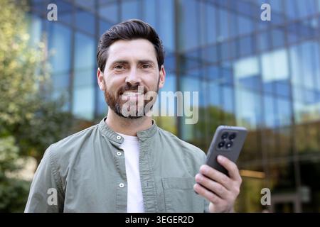 Smiling man with a beard holding a mobile phone, casually dressed and standing in front of a modern glass office building, representing urban lifestyle and connectivity Stock Photo