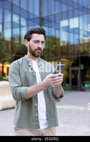 Young man using smartphone and wireless earbud on a downtown street, connecting with mobile apps and online messaging while walking through a modern urban business district Stock Photo