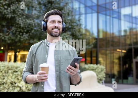 Happy bearded man enjoying a refreshing coffee and listening to music using headphones while strolling outdoors in a modern urban environment, embracing leisure and technology Stock Photo