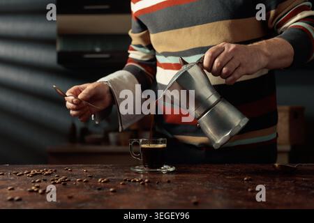 A man in a striped sweater is poured coffee from a geyser coffee maker into a glass cup. It's an authentic vintage Italian coffee maker, a 1950s and 1 Stock Photo