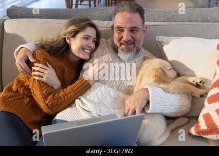 Mature man, mid-aged woman sitting on sofa at home, wearing cable knits, holding laptop and puppy Stock Photo