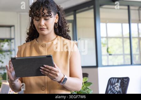 Woman wearing sleeveless tan buttoned top and smartwatch standing in modern office holding tablet Stock Photo