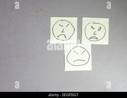 Stick notes feature simple drawings of faces showing emotions. Three notes rest on a grey surface. Each face displays a different expression, including anger and sadness. Stock Photo