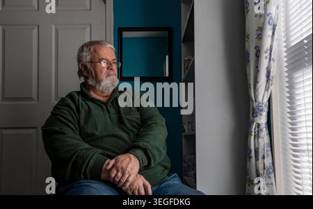 A elderly man with gray hair and glasses sits on a chair beside a window. He looks outside into the daylight. The room has a blue wall and a shelf nearby with items on it. Stock Photo