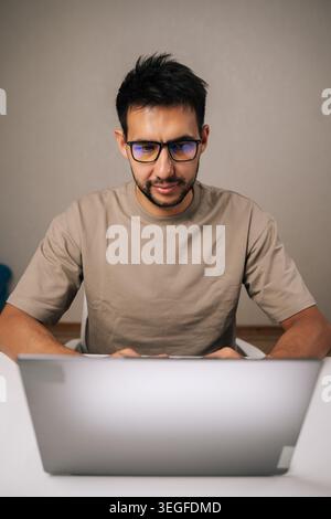 Vertical portrait of bearded young programmer male wearing glasses concentrating on work on laptop, typing on keyboard in remote home office setting Stock Photo