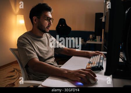 Programmer male concentrating on coding project, desktop screen glowing softly, workspace dimly lit by desk lamp and backlit keyboard during late Stock Photo
