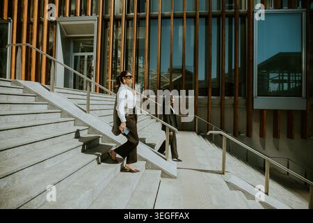 Two business professionals are walking down the steps of a modern building on a sunny afternoon. One person is dressed in white with sunglasses, while Stock Photo