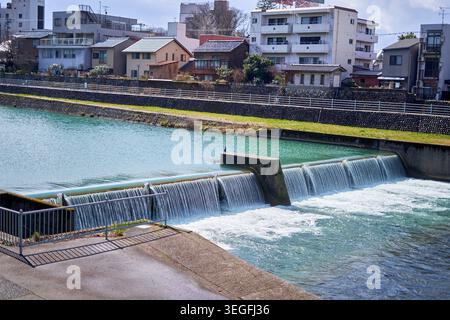 A scenic view of a stepped concrete weir or small dam on a turquoise-colored river flowing through a Japanese residential neighborhood. The structure Stock Photo