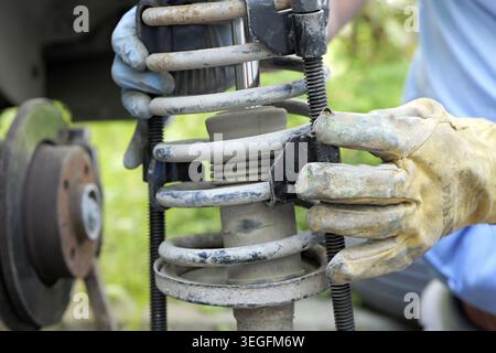 Mechanic's hands in safety gloves using a spring compressor tool during car suspension repair and maintenance in a workshop or outdoor garage environm Stock Photo