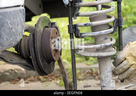 Mechanic's hands in safety gloves compressing a dirty coil spring assembly with a special tool while working on a car's exposed brake disc and suspens Stock Photo