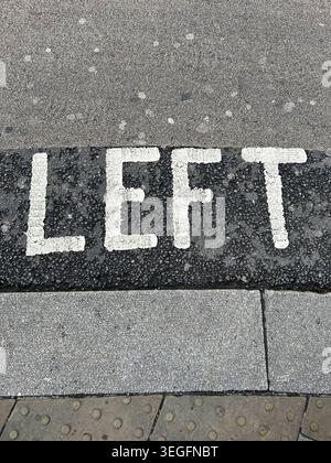 High-angle close-up of the word LEFT painted in white capital letters on a dark road surface next to a stone pavement, used for traffic safety and dir Stock Photo