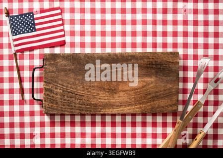 Rustic wooden board with BBQ tools and USA flag on red checkered tablecloth. Top view independence day 4th of July grilling concept. Copy space Stock Photo