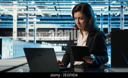 Data center specialist crosschecking settings between laptop and tablet, monitoring industrial systems. Server room computer scientist using devices to oversee network gear performance Stock Photo