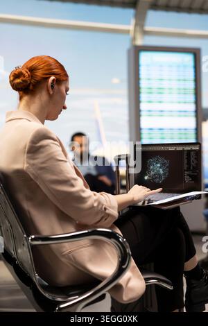Businesswoman using AI powered system on laptop in airport terminal, analyzing big data with machine learning algorithms. Corporate travel concept highlighting smart technology. Stock Photo
