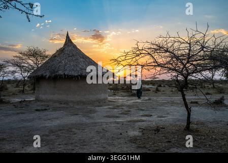 A traditional thatched-roof tukul hut stands against a vibrant golden sunset in Ethiopia. A woman walks near the structure surrounded by acacia trees, Stock Photo