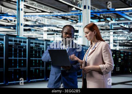 Data center engineering team brainstorming, managing infrastructure systems for uptime and efficiency. Server room teamworking colleagues upgrade enterprise networks hardware using device Stock Photo