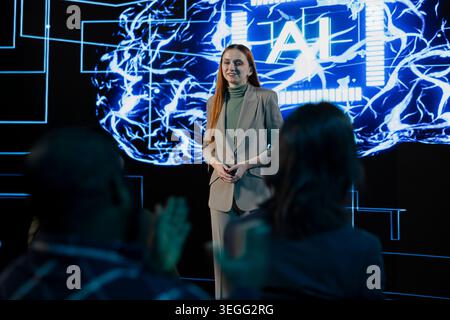 Businesswoman presenter does AI systems presentation with AI graphics, neural networks and deep learning concepts. Business discussion with audience in a high tech conference hall. Stock Photo