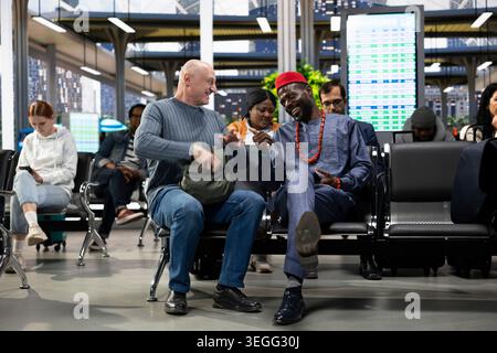 Diverse men becoming friends waiting for daily commute coach, chatting and laughing together in waiting area at bus station. Multiethnic people using the metropolitan commercial service. Stock Photo