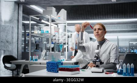 Hematology specialist in lab conducting research on blood sample, taking notes. Epidemiologist in laboratory analyzing sanguine fluid specimen in vial, writing down findings, camera A Stock Photo