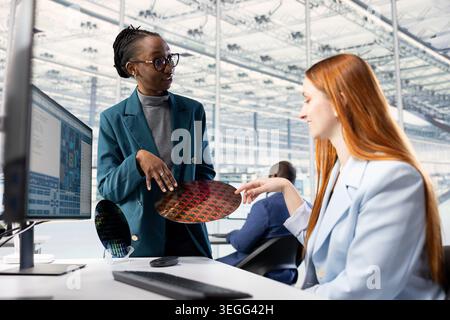 Women looking at silicon wafer, doing quality inspection in microchip factory RD lab. Female engineers examining intricate circuit patterns on semiconductor substrates in office Stock Photo