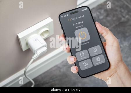 Female hand holding smartphone with smart home application interface for controlling a smart socket on a wall in the background. Concept of home autom Stock Photo