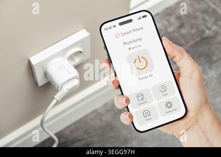 Female hand holding smartphone with smart home application interface for controlling a smart socket on a wall in the background. Concept of home autom Stock Photo