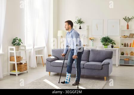 Young man with broken leg after accident using his crutches standing in living room at home. Stock Photo