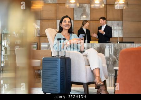 Smiling female hotel guest sitting in reception using smartphone Stock Photo