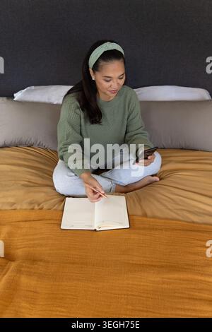 Mid adult Asian woman sitting on bed wearing green sweater holding smartphone pencil notebook Stock Photo