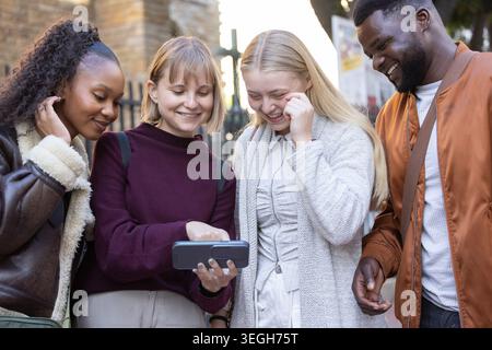 Diverse friends leaning in, pointing at smartphone on city sidewalk by brick building wearing coats Stock Photo