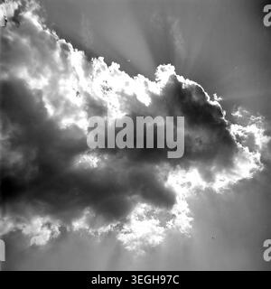 Gurzuf, 1960s, a dramatic view of the sky where a large, dark cumulus cloud partially obscures the bright sun. Distinct sunbeams, also known as crepuscular rays, pierce through the atmospheric haze and radiate outward across the firmament. This striking meteorological phenomenon creates a powerful contrast between deep shadows and glowing edges in the heavens. The vintage black and white frame captures the raw beauty and majestic power of natural weather over the coast. A quiet glimpse into peaceful life before the war. Stock Photo