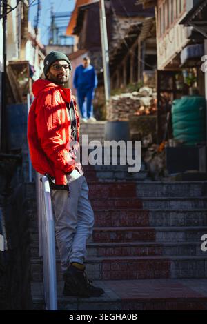 A young man wearing a red jacket and beanie stands on stone steps in a rustic village setting, Chakrata, Uttarakhand, India. Stock Photo