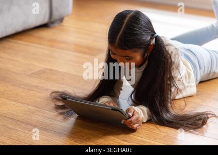 Female child lying on stomach using tablet on hardwood floor at home, wearing light sweater Stock Photo