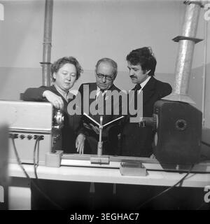Sloviansk, late 1970s - early 1980s. Inside a brightly lit laboratory or factory workspace, three professionals are deeply engaged in analyzing technical data. A woman and two men in formal business attire and suits stand closely together, attentively reading an open manual or logbook next to complex optical and electronic testing equipment. The serious atmosphere highlights their dedication to scientific research, quality control, and industrial progress. This candid moment provides a fascinating glimpse into professional life and peace in Donbas. Stock Photo