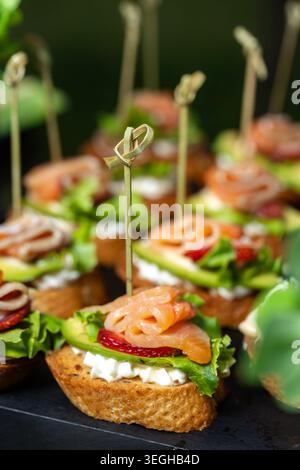 A vertical close-up shot displays elegant catering bruschettas topped with salted salmon, fresh avocado, strawberry slices, and cream cheese. Stock Photo