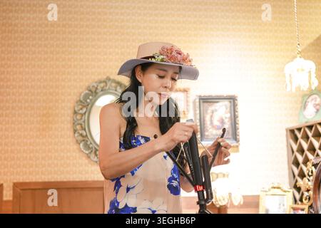 Woman wears floral hat and dress, focused on camera. Her expression shows quiet concentration as she adjusts settings. Soft lighting highlights warm, Stock Photo