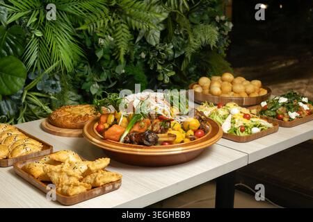 A light wooden table displays assorted platters featuring roasted meat, pastries, and fresh salads against a lush green wall. Stock Photo