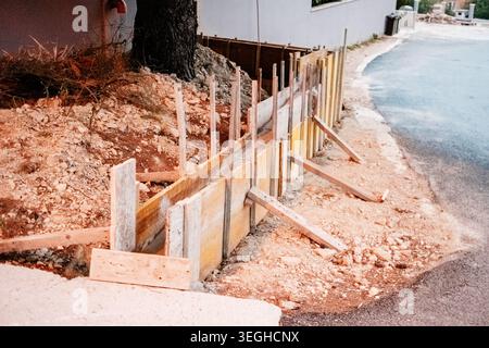 Temporary wooden formwork is precisely set for pouring a small concrete retaining wall along a dusty construction site next to a paved road. Stock Photo