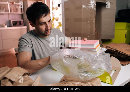 Young man in casual clothes packs books using bubble wrap and cardboard boxes. Domestic scene of relocating or organizing personal belongings in Stock Photo