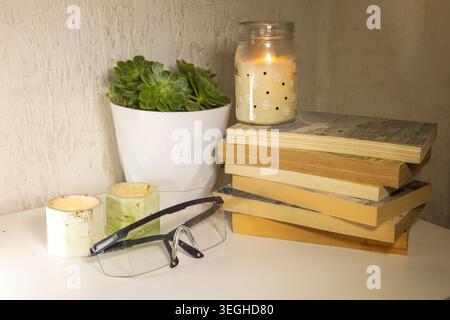 A white desk features a stack of books, a lit jar candle, two decorative holders, a potted succulent, and clear safety glasses. Stock Photo