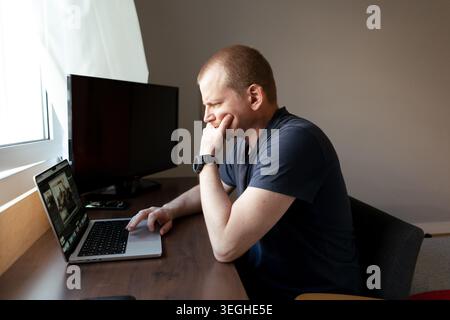 Man focusing on a laptop computer screen, holding his chin while working from an office desk Stock Photo