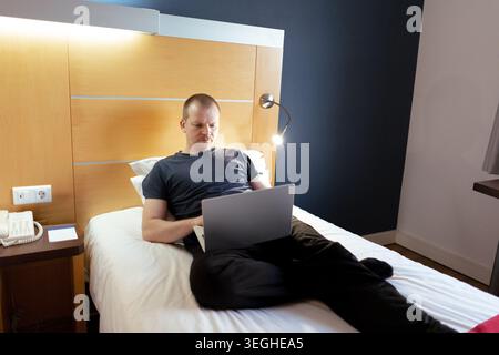 Man lying in bed using a laptop for remote work in a hotel room Stock Photo