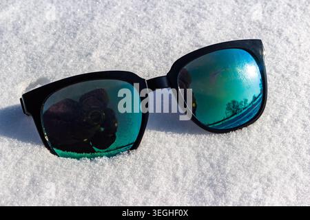 Stylish black sunglasses with vibrant mirrored lenses lie on sparkling snow, reflecting a bright winter scene and the photographer Stock Photo