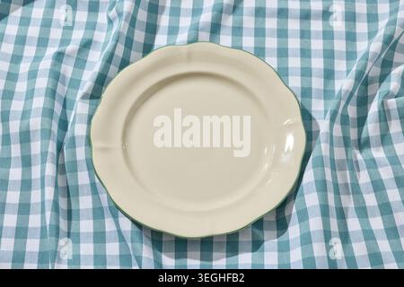 Overhead flat lay shot of an empty minimalist white ceramic plate centered on a rustic, wrinkled blue and white checkered linen tablecloth. Captured i Stock Photo