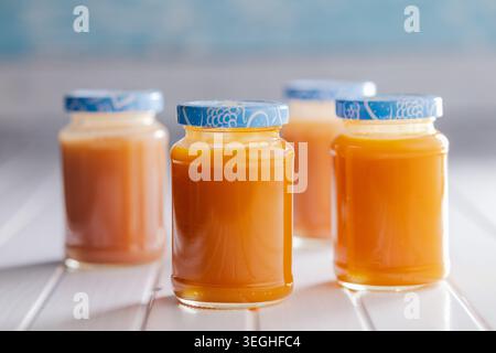 Four glass jars filled with smooth orange fruit puree, sealed with patterned lids. Warm, homemade baby food or jam jars arranged on a bright white tab Stock Photo