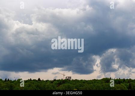 Dramatic overcast spring sky filled with heavy storm clouds hanging above a lush green forest canopy featuring a distant construction site with a cran Stock Photo